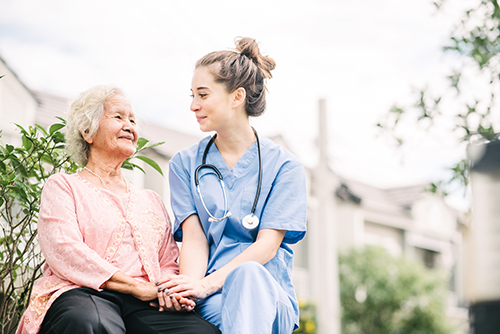 Young nurse wearing stehtoscope sitting outside with elderly woman holding her hand