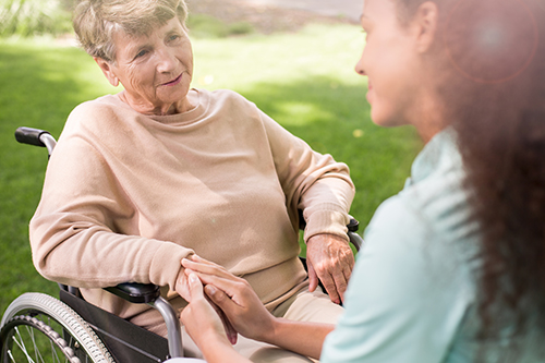 Nurse in teal scrubs knelt down next to elderly woman in wheelchair holding her hand