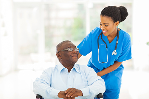Nurse in blue scrubs standing behind elderly man in wheelchair both smiling at each other